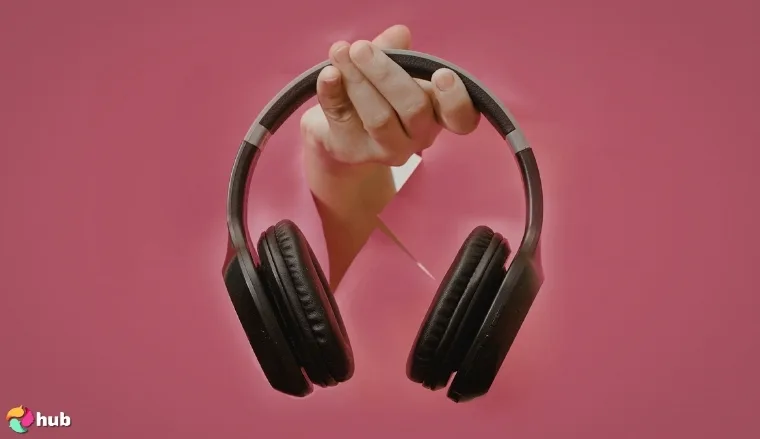 A hand holding black wireless headphones against a pink background