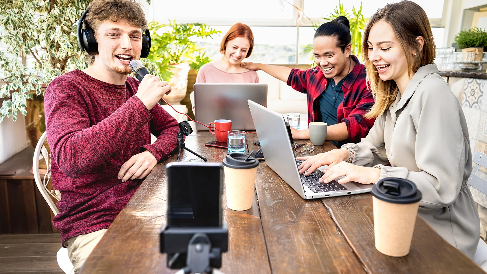 Group of people recording a podcast or video with a microphone, smartphones, and laptops in a bright room