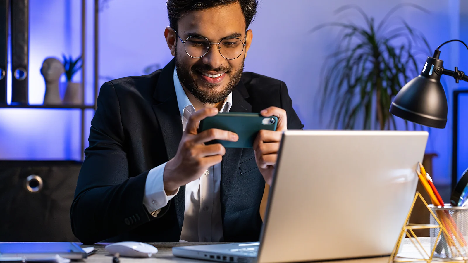 Man smiling while testing a mobile game on his smartphone at a desk with a laptop