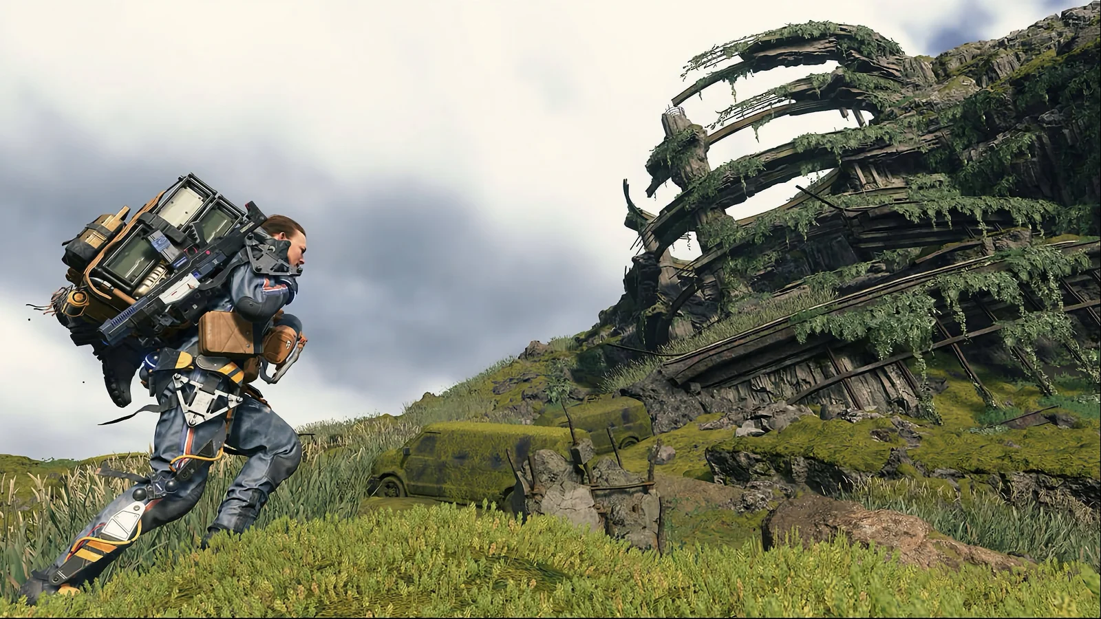 Death Stranding – Melhor imersão na travessia – Uma perspetiva na terceira pessoa de Sam Porter Bridges, vestido com um fato futurista e a carregar uma pilha enorme e pesada de contentores nas costas, enquanto caminha por uma encosta verdejante e coberta de relva em direção a uma estrutura metálica colossal em ruínas, coberta de musgo