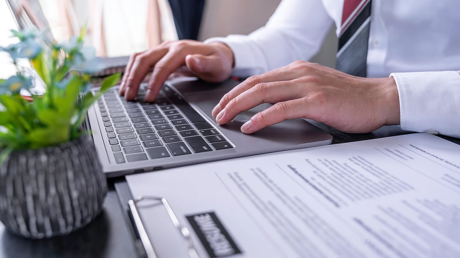 Business professional typing on a laptop next to a printed resume on a clipboard