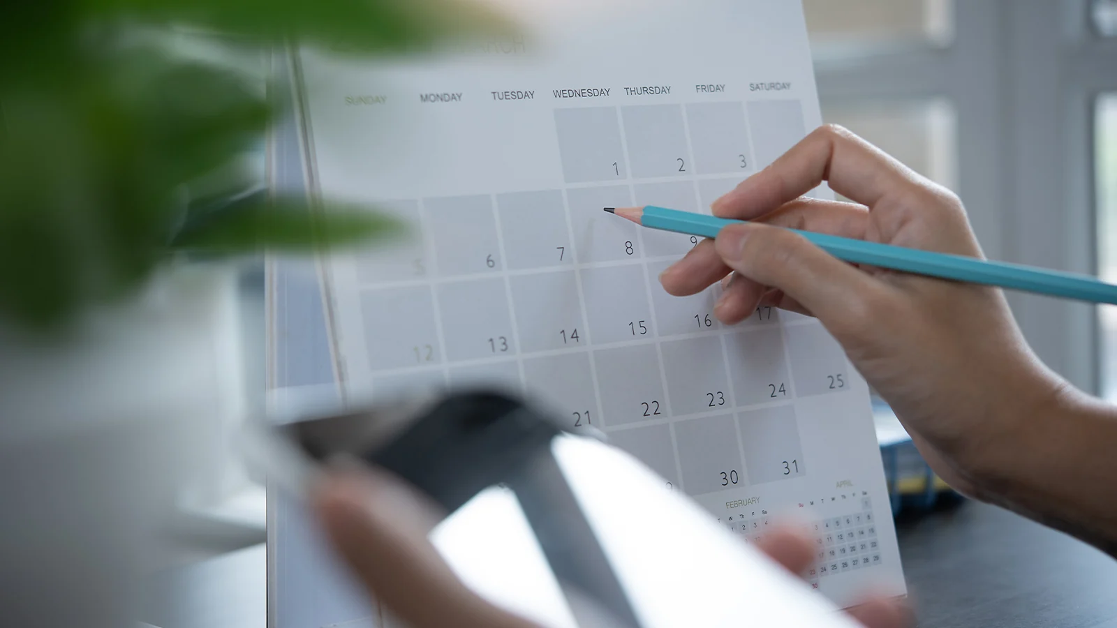 Person using a pencil and smartphone to plan rotating nursing shifts on a desk calendar